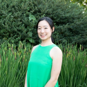 A woman stands with her arms at her sides smiling at the camera. She is sanding in front of bright green cattails and green trees are behind her. She is wearing a green dress and has brown hair. She is Korean and looks to be in her 20's or 30s.